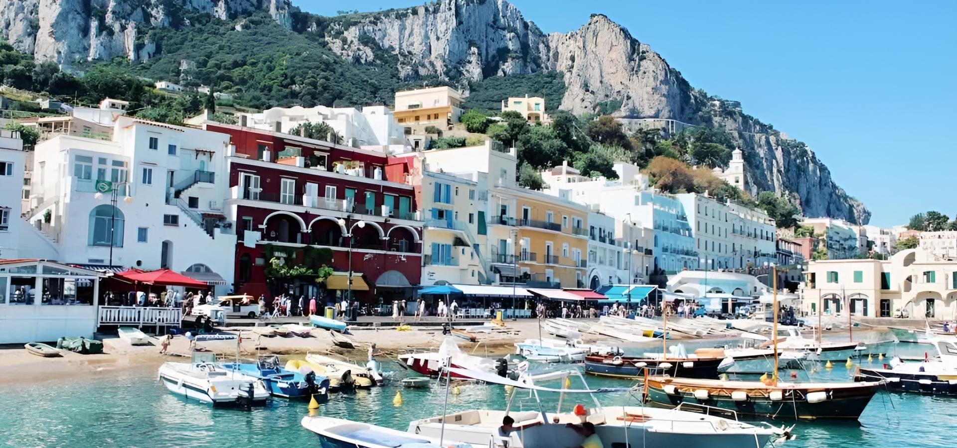 Porticciolo colorato con barche, case vivaci e montagne imponenti sotto un cielo azzurro.