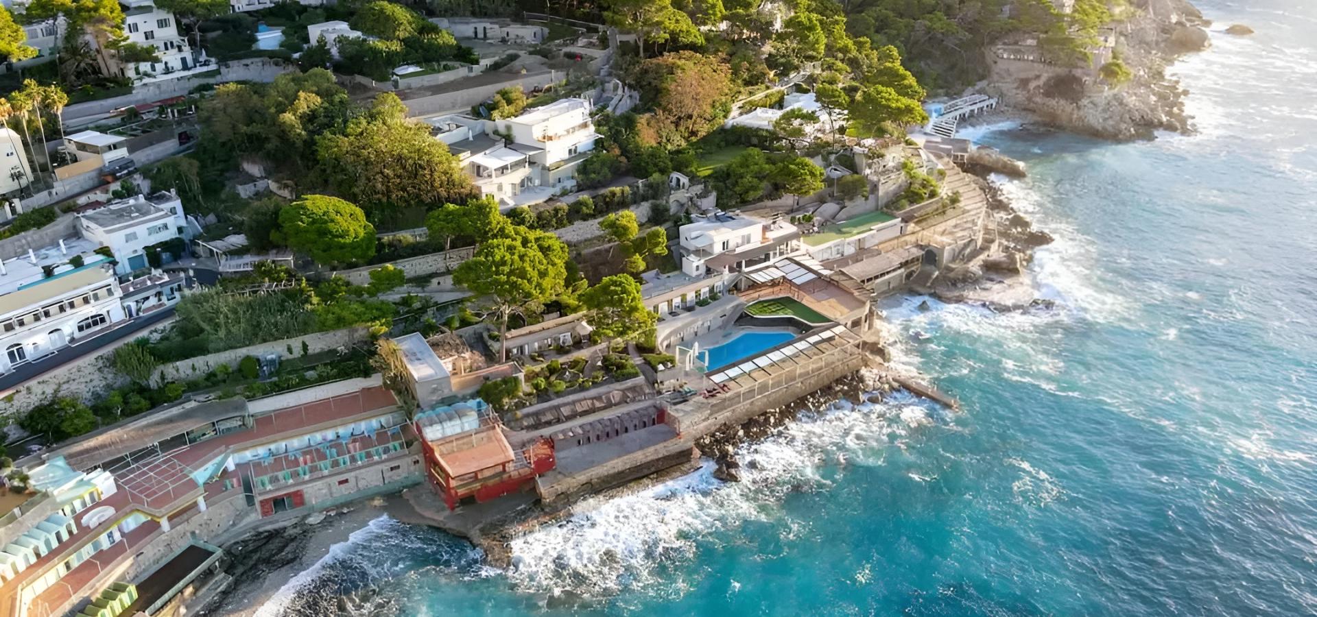 Vista aerea di una costa rocciosa con ville lussuose e mare azzurro scintillante.