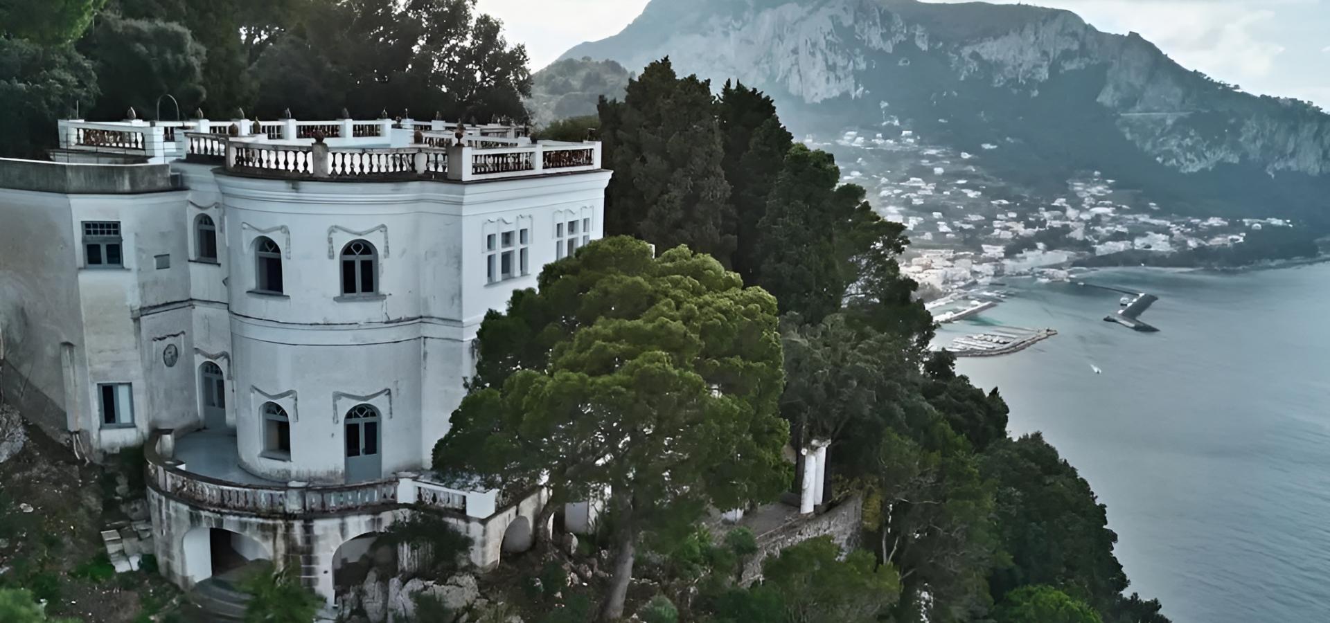 Villa bianca su scogliera, circondata da alberi, con vista mozzafiato sul mare e montagne.