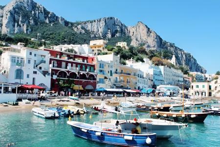 Porticciolo colorato con barche, case vivaci e montagne imponenti sotto un cielo azzurro.