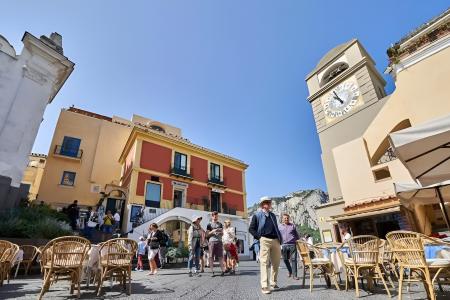 Piazza vivace con orologio, cielo azzurro, tavolini all'aperto e persone passeggianti.