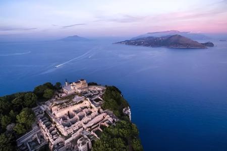 Vista aerea di rovine antiche su un'isola al tramonto, circondate dal mare blu.