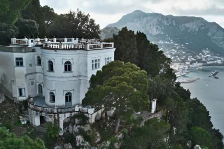 Villa bianca su scogliera, circondata da alberi, con vista mozzafiato sul mare e montagne.